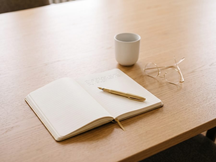 An open notebook with a gold fountain pen, a coffee cup and reading glasses on an oak seminar table