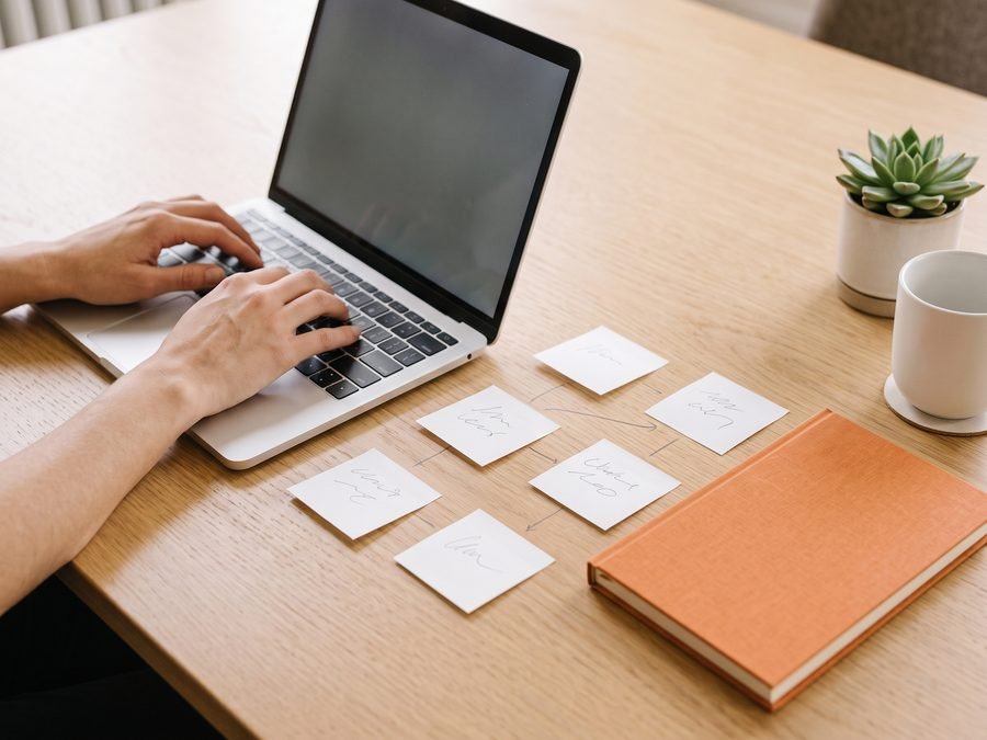 Hands typing on a laptop beside a flow of sticky notes and an orange notebook